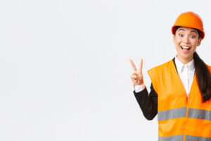 Happy smiling asian female engineer, architect in safety clothins and helmet, showing peace kawaii sign and looking upbeat, company winning tender on construction and building, white background